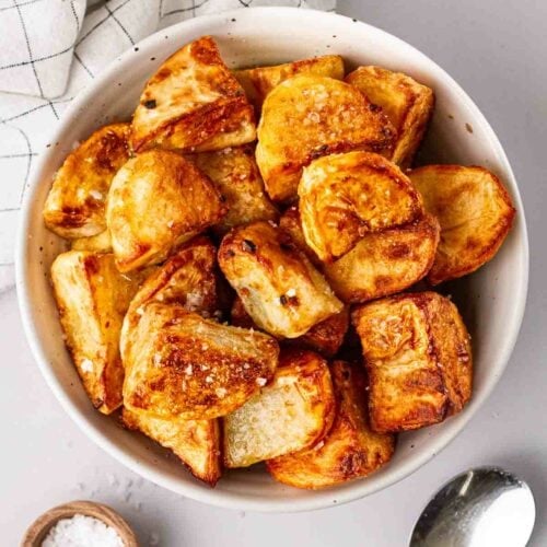 A bowl filled with roast potatoes made in the air fryer, on a white table with a white cloth to the side, a small pinch pot of salt and a spoon.