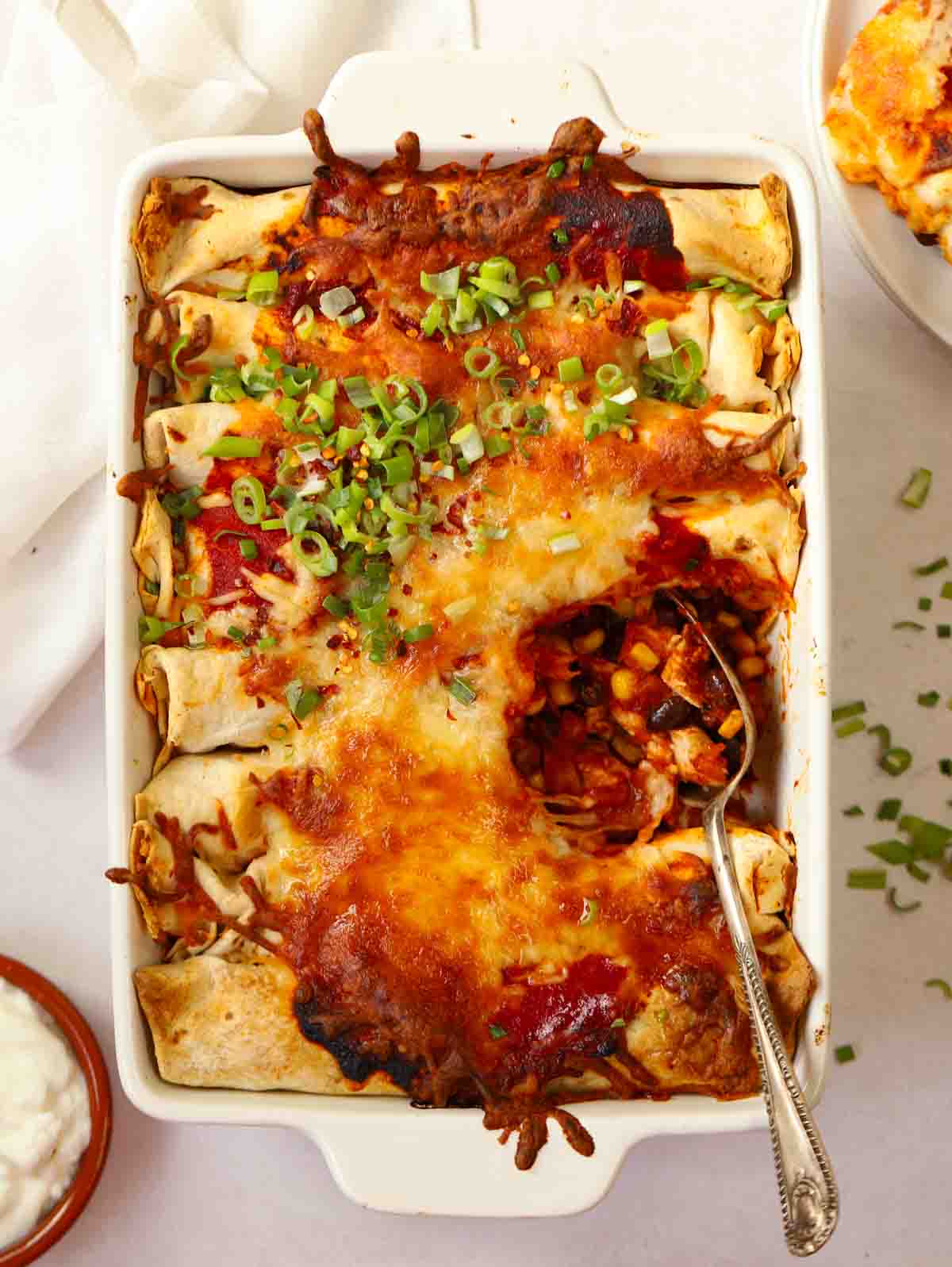 A white ovenproof dish filled with Chicken Enchiladas with a spoonful missing on a white table cloth.