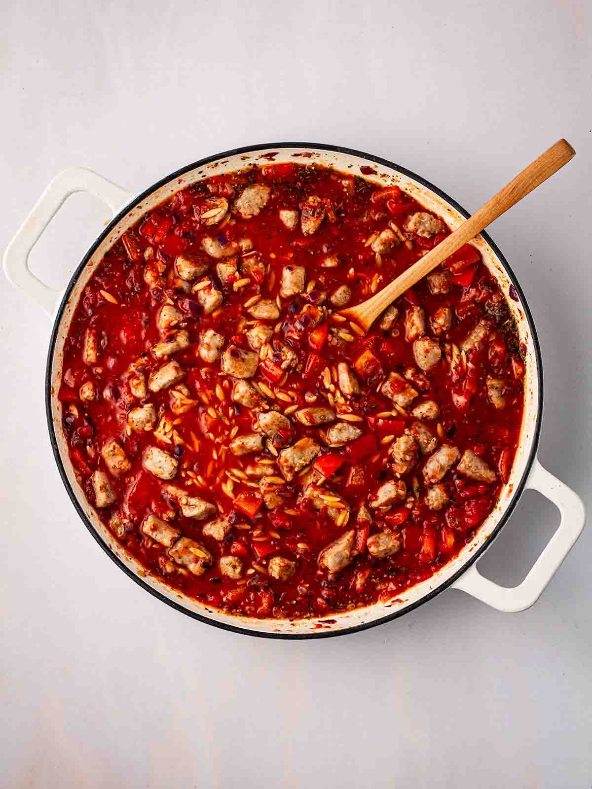 Chunks of sausage and red pepper in a pan with orzo and tomato sauce, with a wooden spoon.