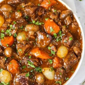 A big bowl of beef bourguignon on a white surface, ready to eat. Filled with meat, onions, carrots in a rich gravy.