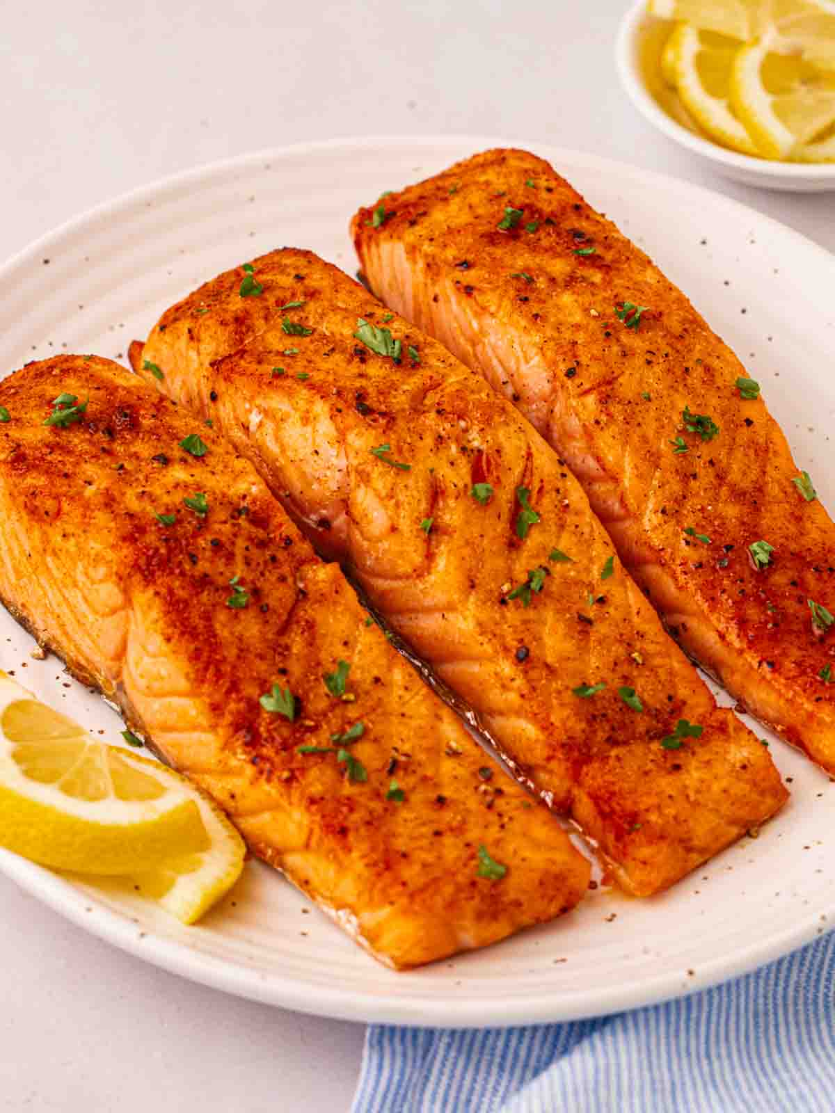 Three cooked air fryer salmon fillets on a white plate with lemon wedges on the side, sprinkled with parsley.