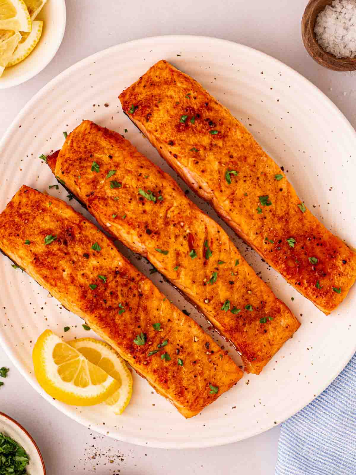 A bird's eye view of three cooked salmon fillets on a white plate with a piece of lemon to the side, as well as a salt pinch pot.