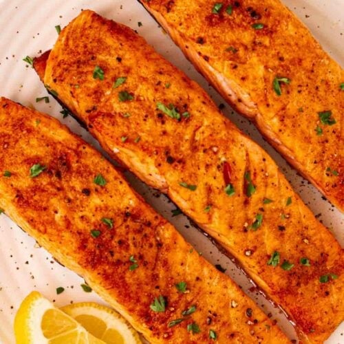 Three salmon fillets that have been cooked in an air fryer on a white plate with lemon slices.