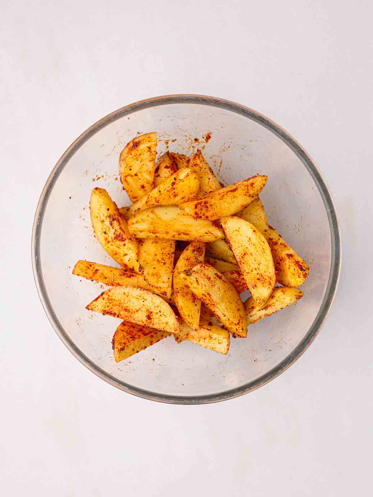 A glass bowl of raw seasoned potato wedges.