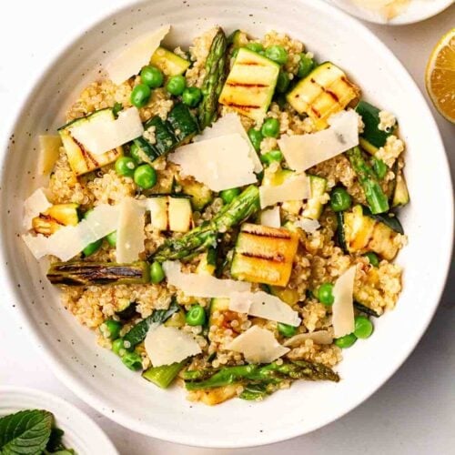 A white counter with a bowl of Quinoa Salad, lemon at the side, with bowls of mint, parmesan and salt and pepper.