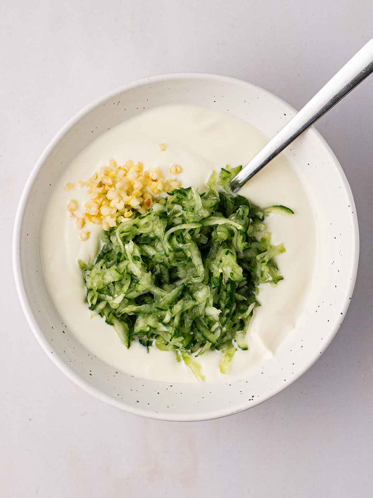 A white bowl with a tzatziki dressing about to the mixed together for the side dish to Salmon Burgers.