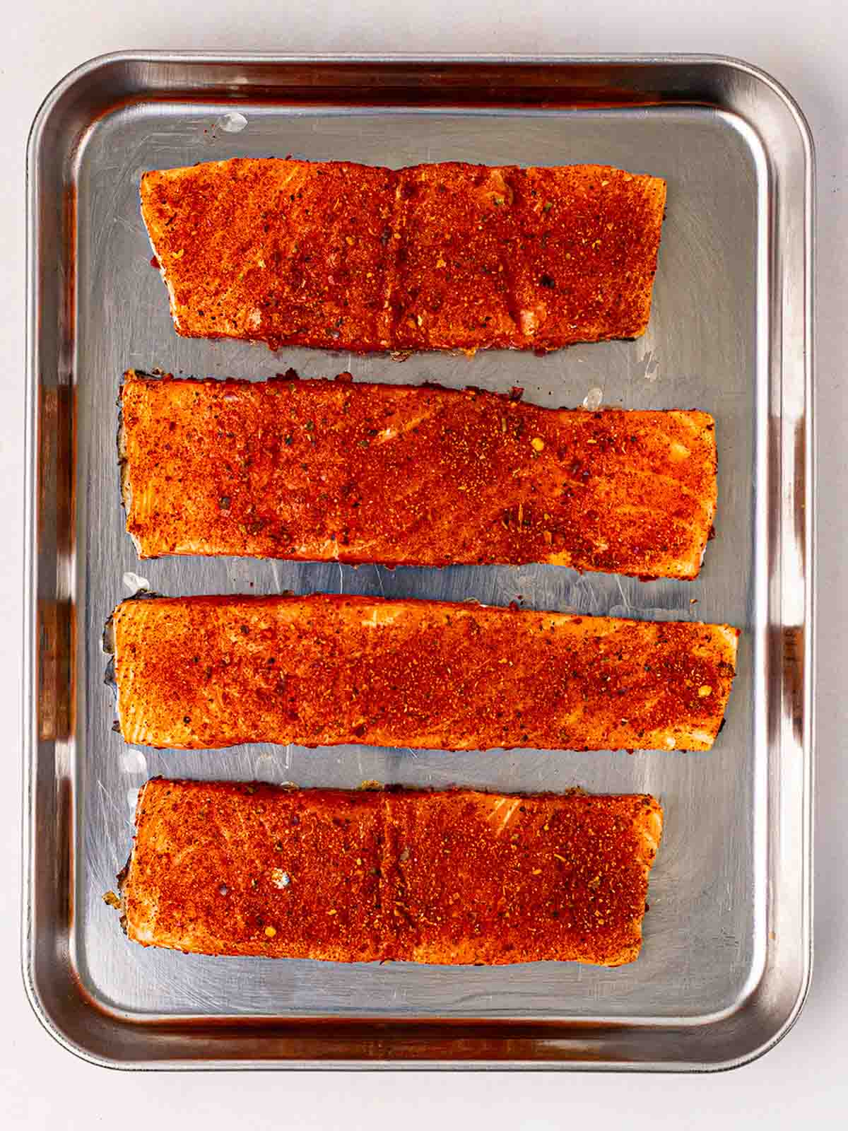 Four salmon fillets covered with cajun spice on a metal baking tray, for step 1 in the recipe for Salmon Tacos.