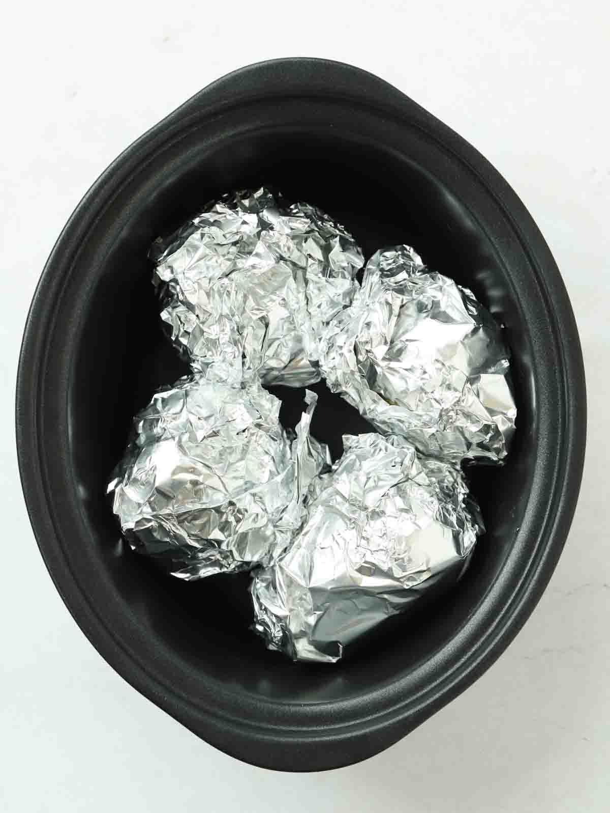 Four potatoes wrapped in foil in a slow cooker pan.