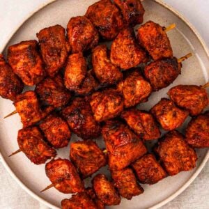 Five skewers with chunks of tandoor chicken on, laying on a plate.