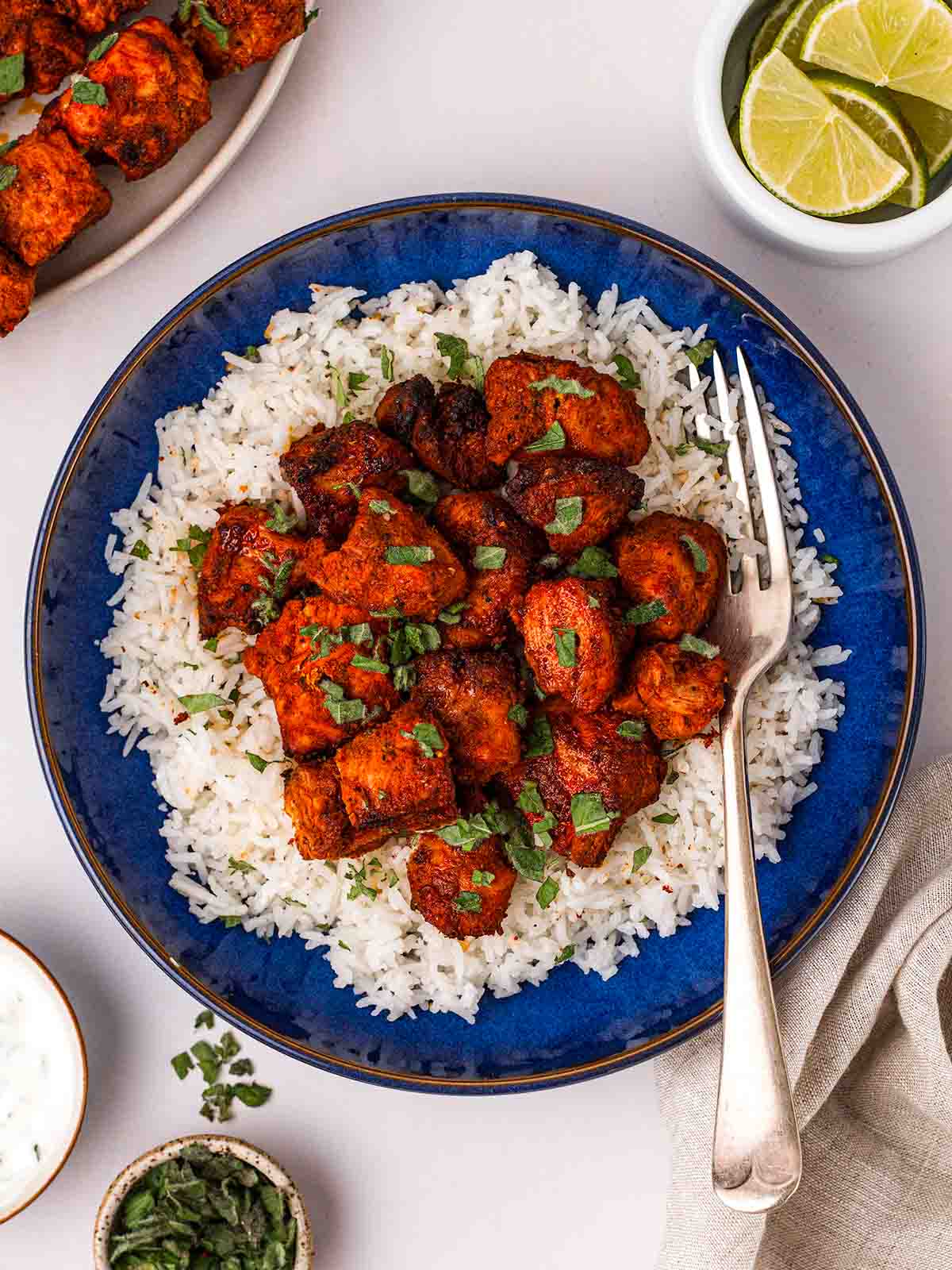 A blue plate with rice and tandoori chicken chunks with a fork and surrounded by lime, coriander and other small pots.