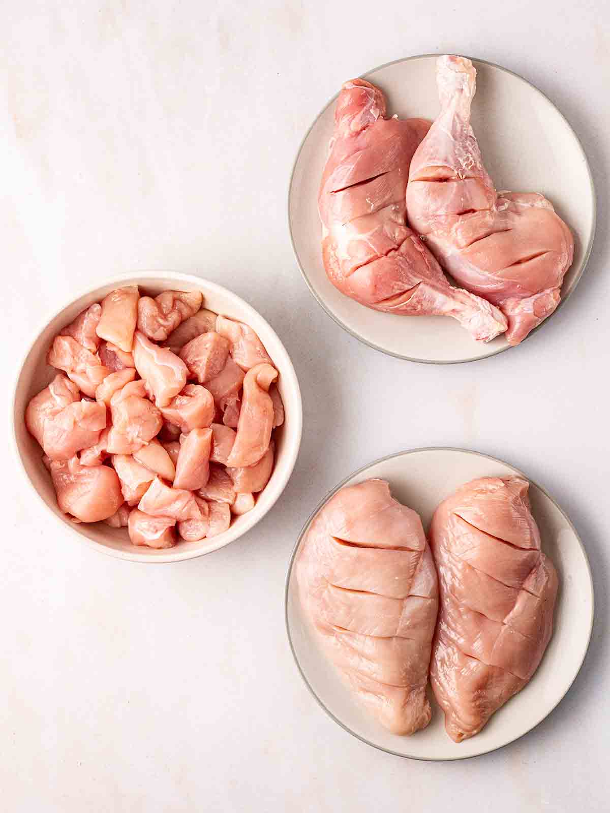 Three plates of raw chicken. One with a skinless chicken leg, one with diced chicken breast, and another with two scored chicken breasts for the recipe Tandoori Chicken.
