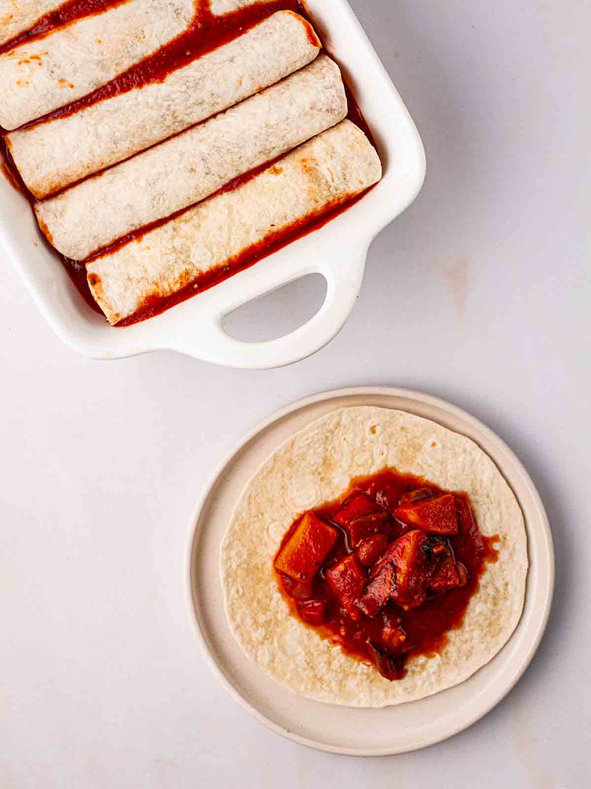 Rolled enchiladas in an oven dish with an unrolled wrap on a plate to show the vegetable filling for step 4 of the recipe for Vegetarian Enchiladas.