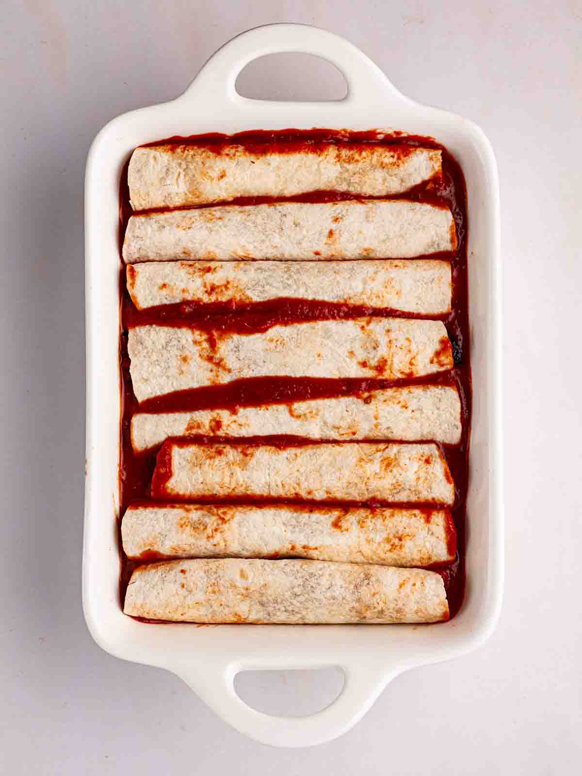 Rolled Vegetarian Enchiladas in a white oven dish for for step 5 of the recipe for Vegetarian Enchiladas.