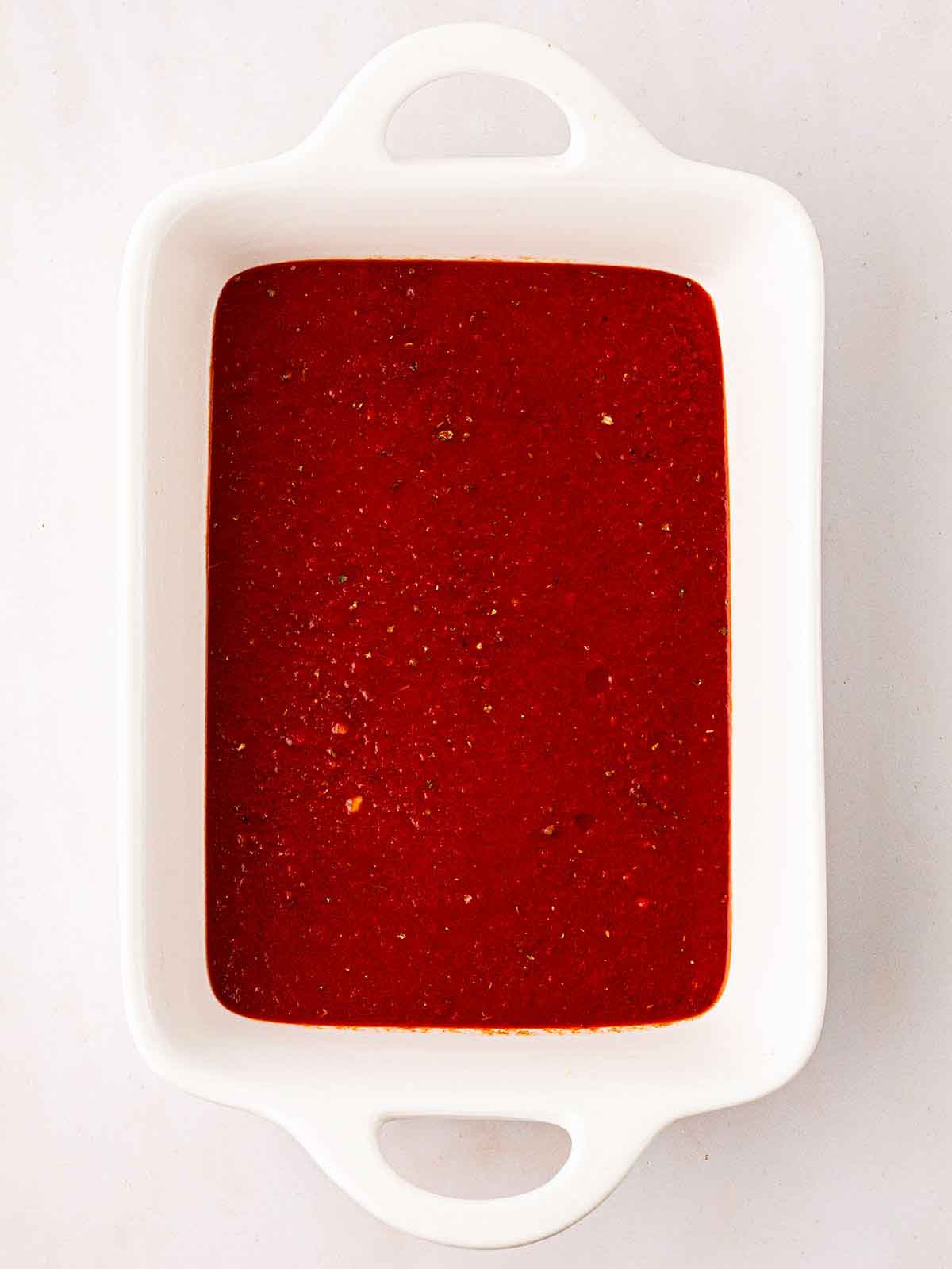 An oven dish with tomato sauce in for for step 3 of the recipe for Vegetarian Enchiladas.