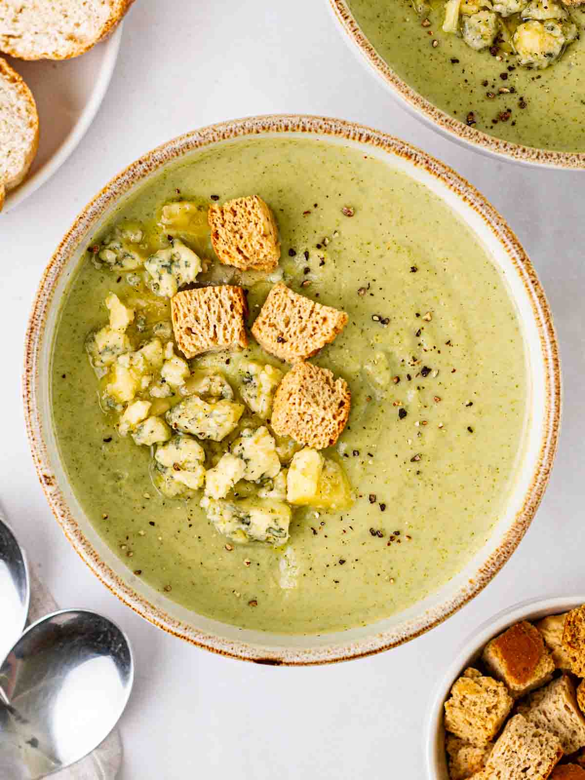 A bowl of Broccoli and Stilton Soup topped with cheese and croutons on a table with soup, bread and spoons.