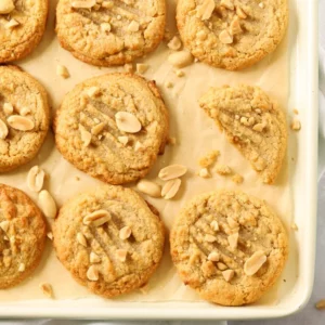 A tray of just baked Peanut Butter Cookies on a lined baking tray with chopped peanuts sprinkled over the top.