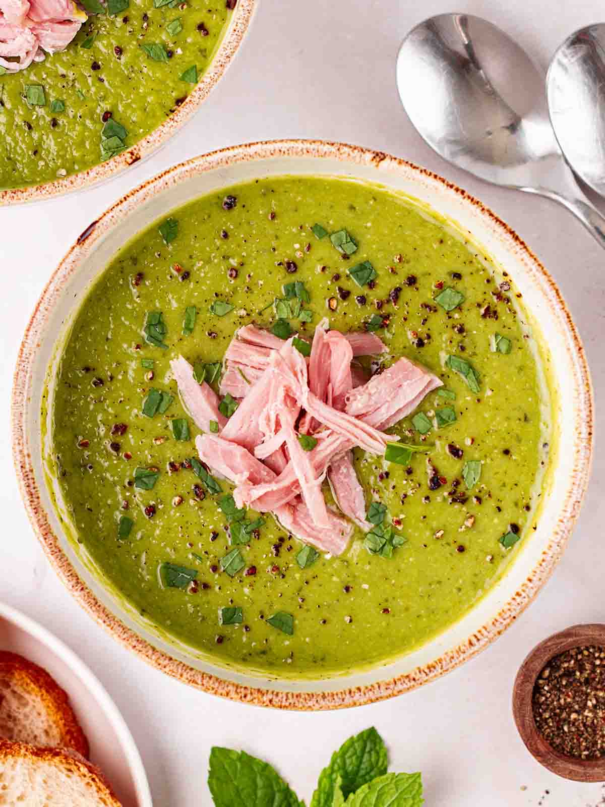 A white counter with bowls of cooked Pea and Ham Soup, with spoons, ready to eat.