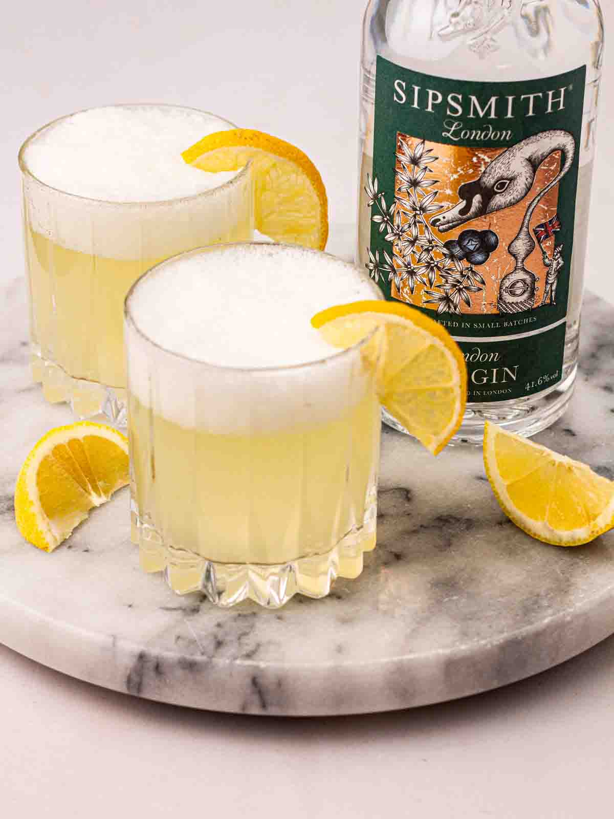 Two glasses of Gin Fizz cocktail with slices of lemon on a marble board with a bottle of Sipsmith gin in the background.