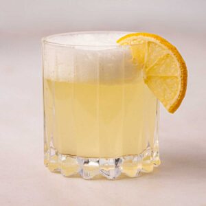 A glass filled with Gin Fizz, with frothy egg white on top and a slice of lemon.