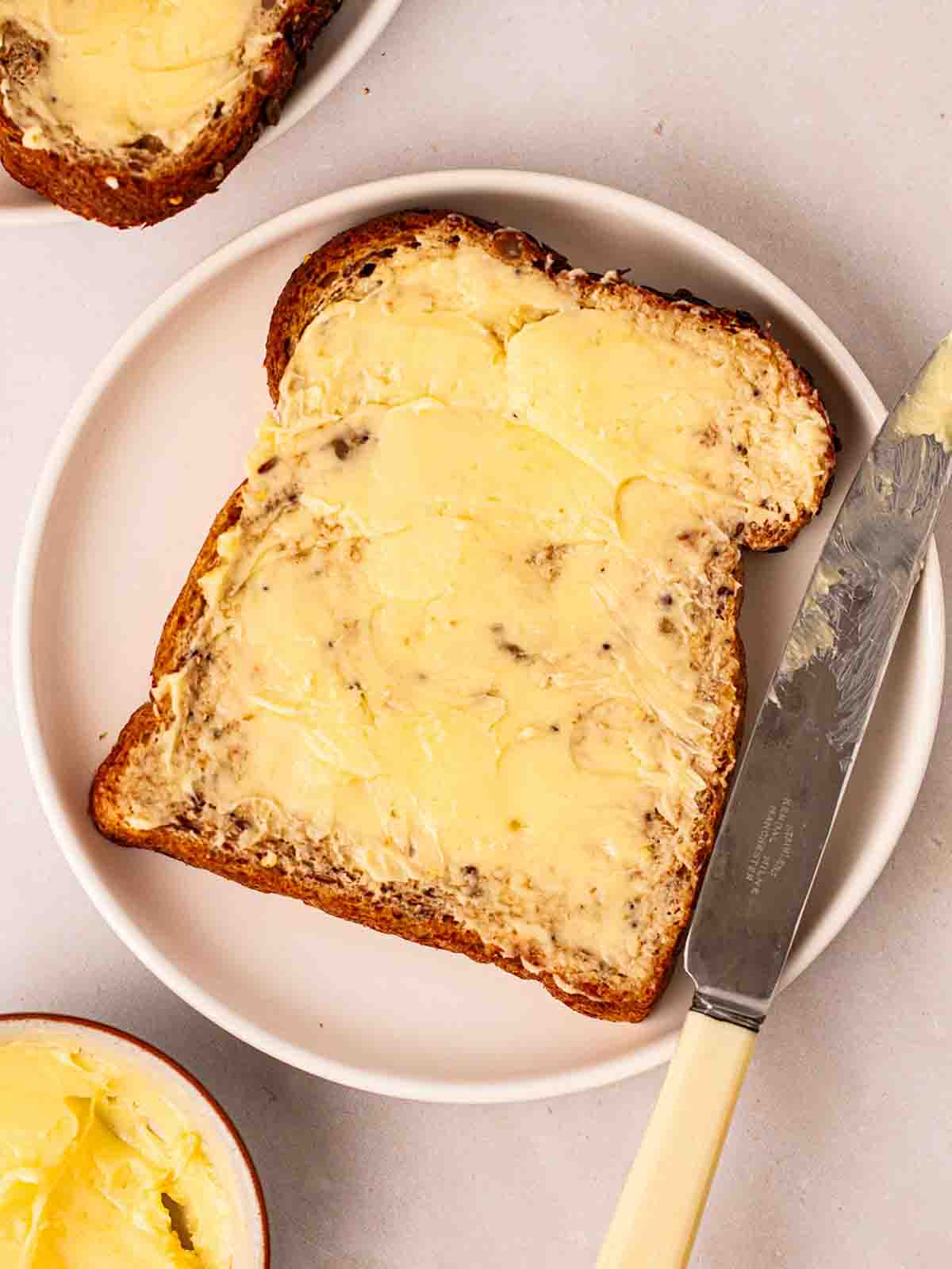 Homemade butter slathered thickly onto a slice of brown bread, on a white plate with a knife to the side.