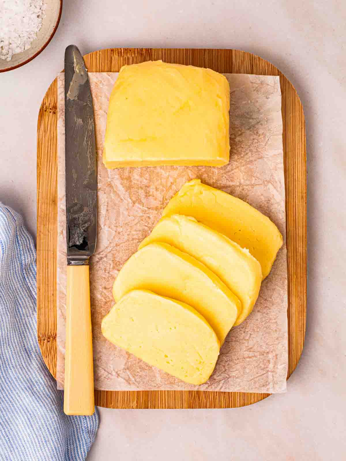 A wooden chopping board with a slab of homemade butter, half of it sliced, with a knife to the side and a layer of baking parchment paper underneath.