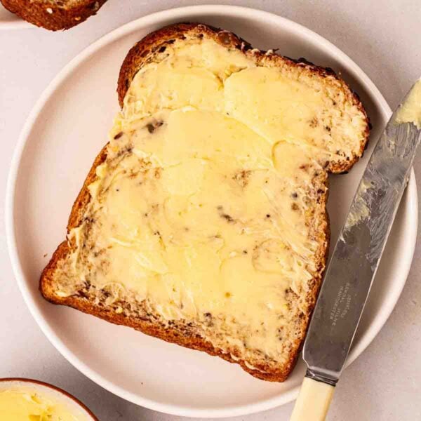 A slice of bread on a plate, with thickly spreaded homemade butter on, and a butter knife next to it.
