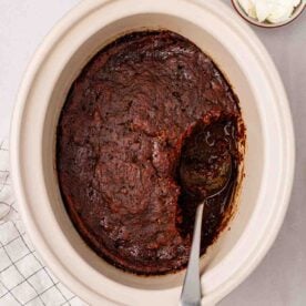 A slow cooker with cooked Sticky Toffee Pudding inside, with a sauce under the sponge. A spoon to serve up.