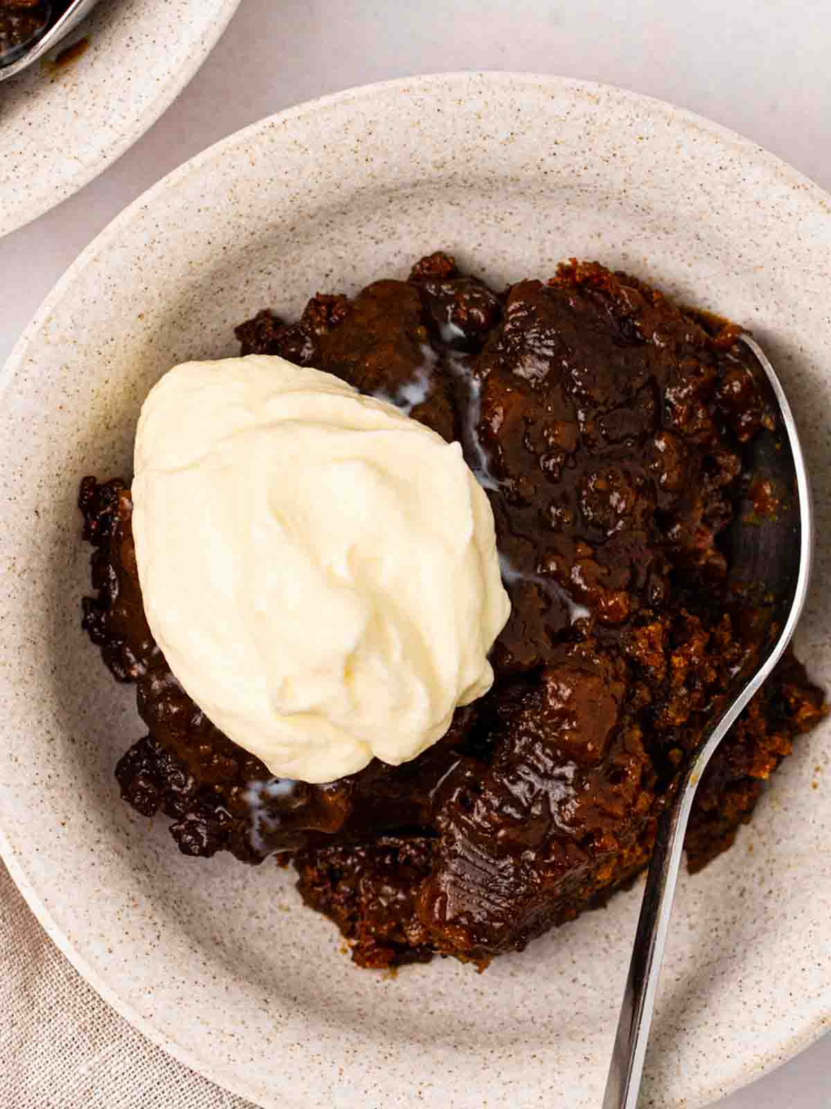 A portion of Sticky Toffee Pudding made in the slow cooker in a bowl with a spoon and dollop of ice cream.