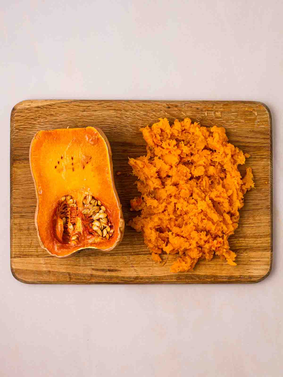A halved butternut squash on a chopping board. One side is intact, the other half is mashed.