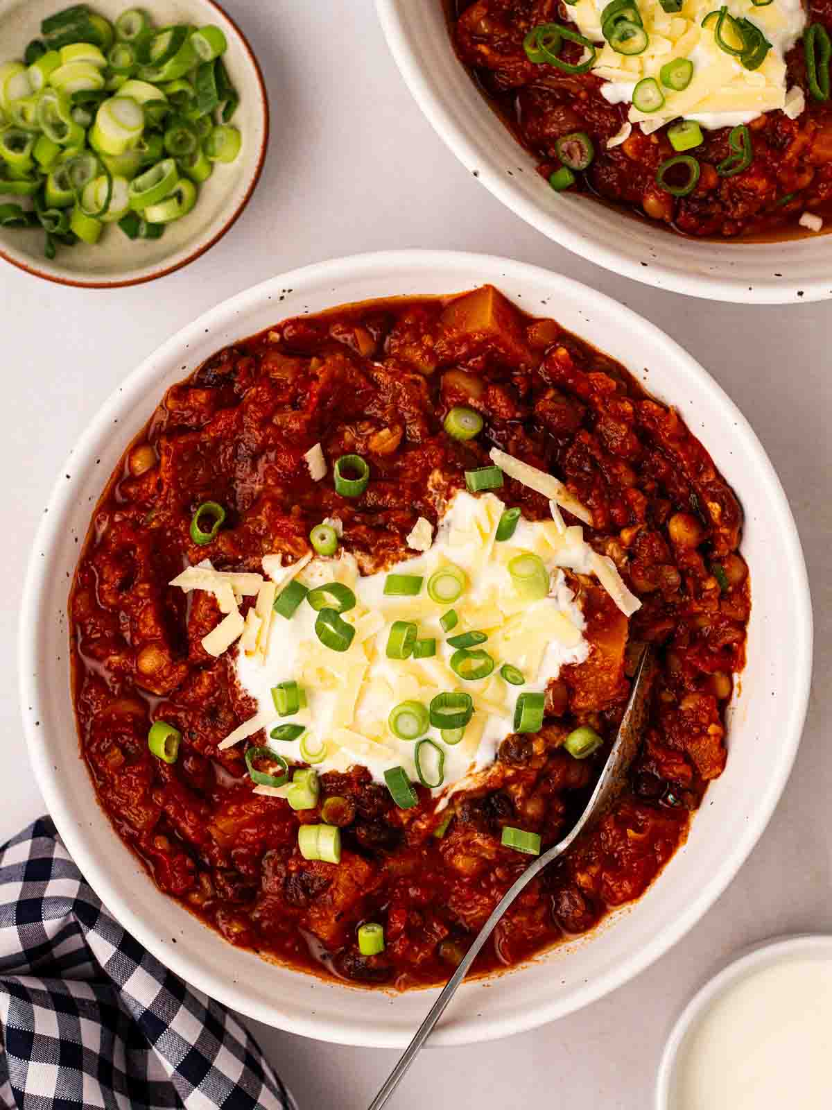 Two bowls filled with vegetarian chilli, topped with sour cream and spring onions, with a spoon.