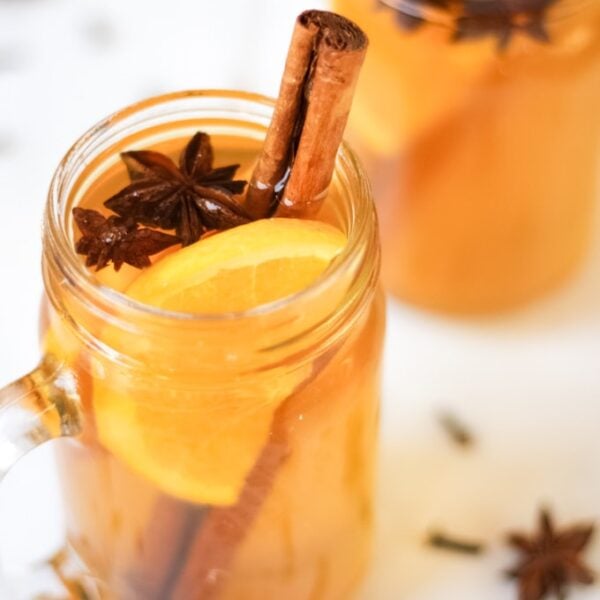 A close up of a glass jar of Mulled Cider, garnished with cinnamon stick, star anise and a slice of orange.