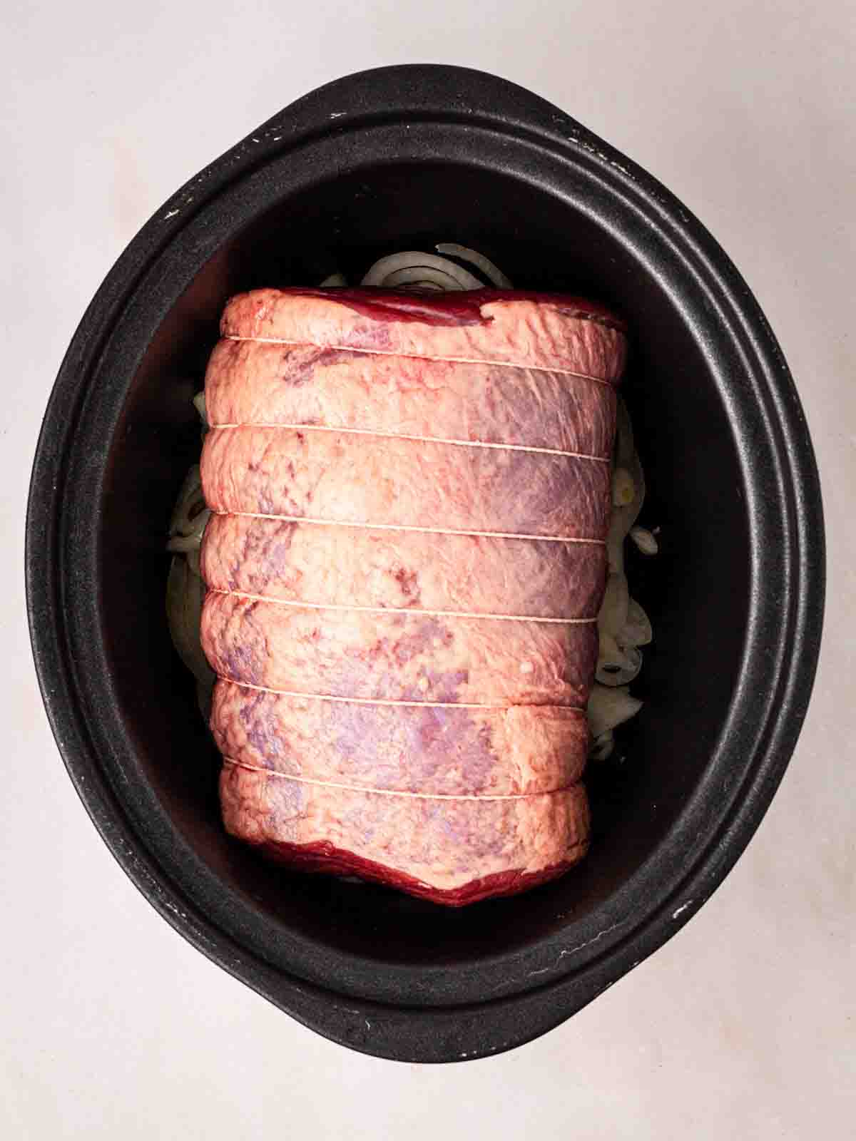 A piece of beef brisket laid on a bed of sliced onions in a large black slow cooker.