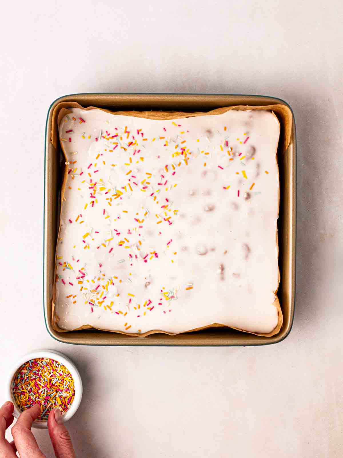 A hand sprinkling over sprinkles on a cake with white icing.