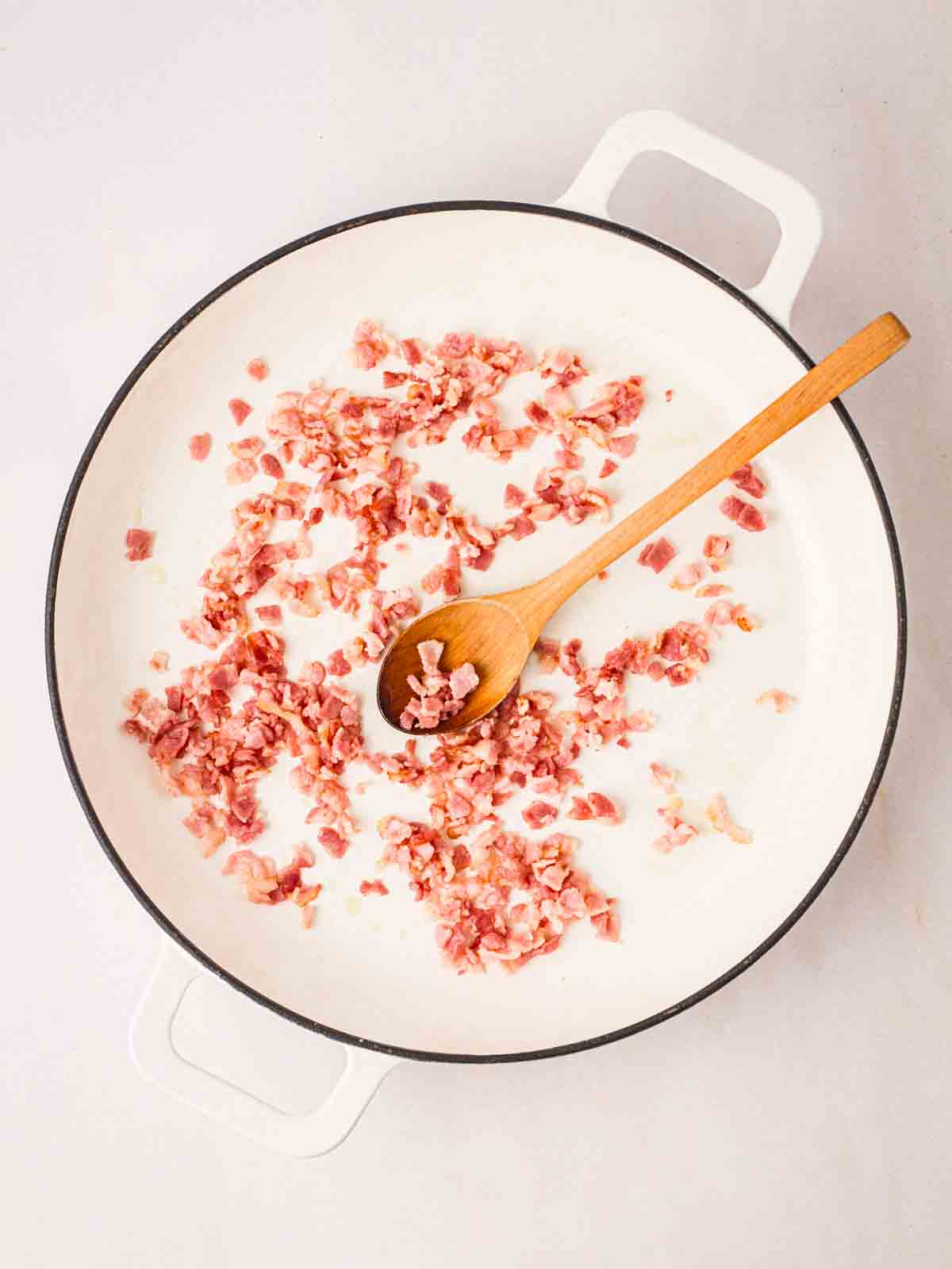 Chopped up pancetta pieces frying in a white pan with a wooden spoon in for step in the recipe for Spaghetti Bolognese.