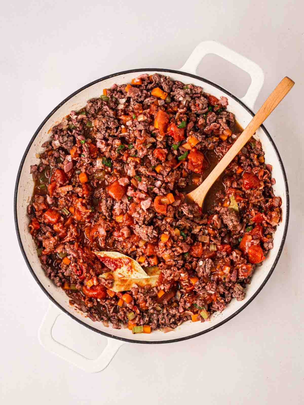 Mince with a tomato sauce, chopped vegetables and herbs in a white pan with a wooden spoon stirring the sauce, for step 5 in the recipe for Spaghetti Bolognese.