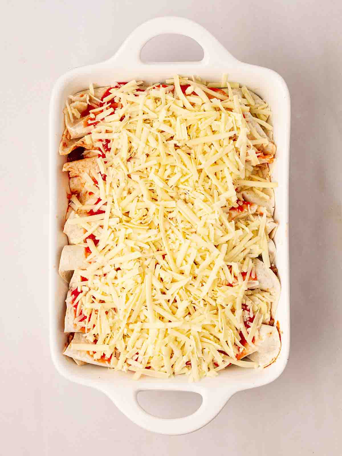 An oven dish with rolled tortillas and sprinkled grated cheese on top for step 6 in the recipe for Beef Enchiladas.