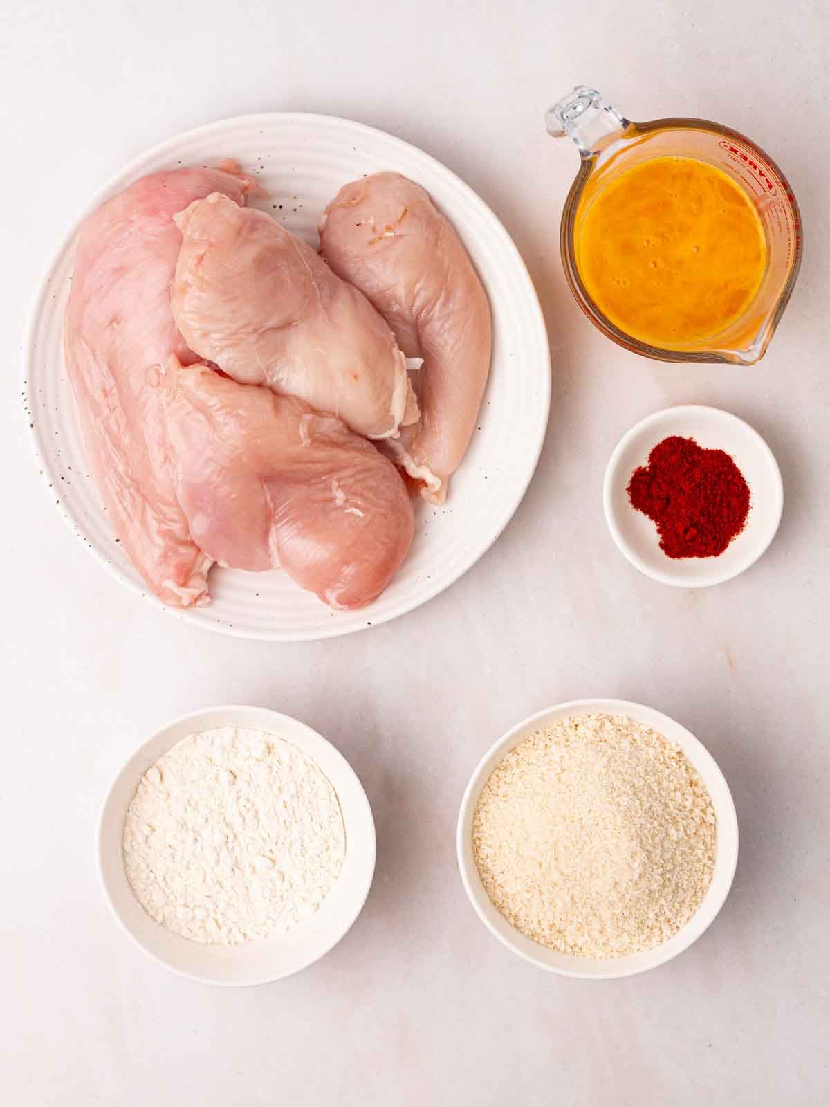 Raw ingredients laid out on a white counter, including chicken breast, spices and breadcrumbs.