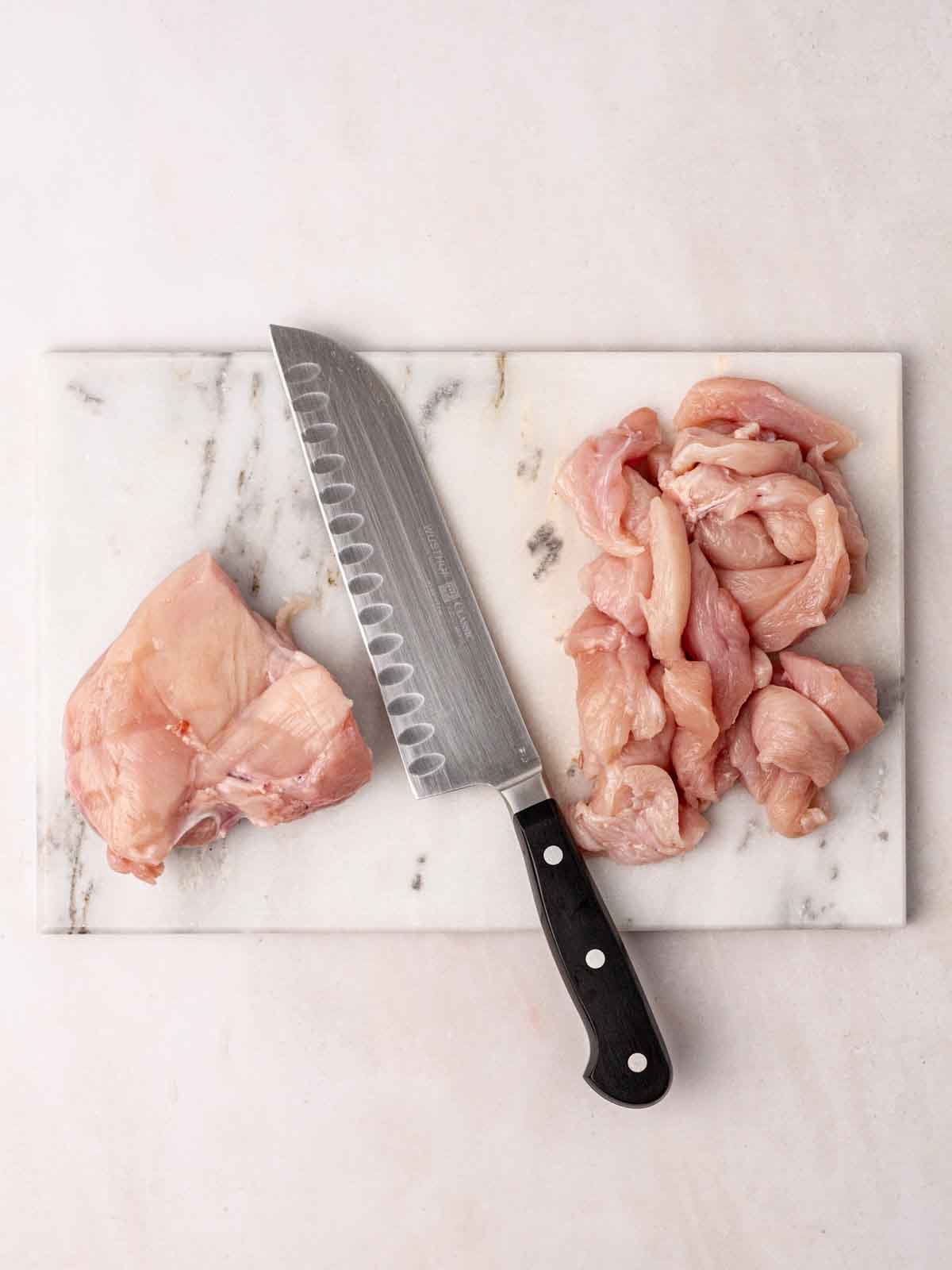 A marble chopping board with a knife and raw chicken breast, with half already cut into bitesized pieces.
