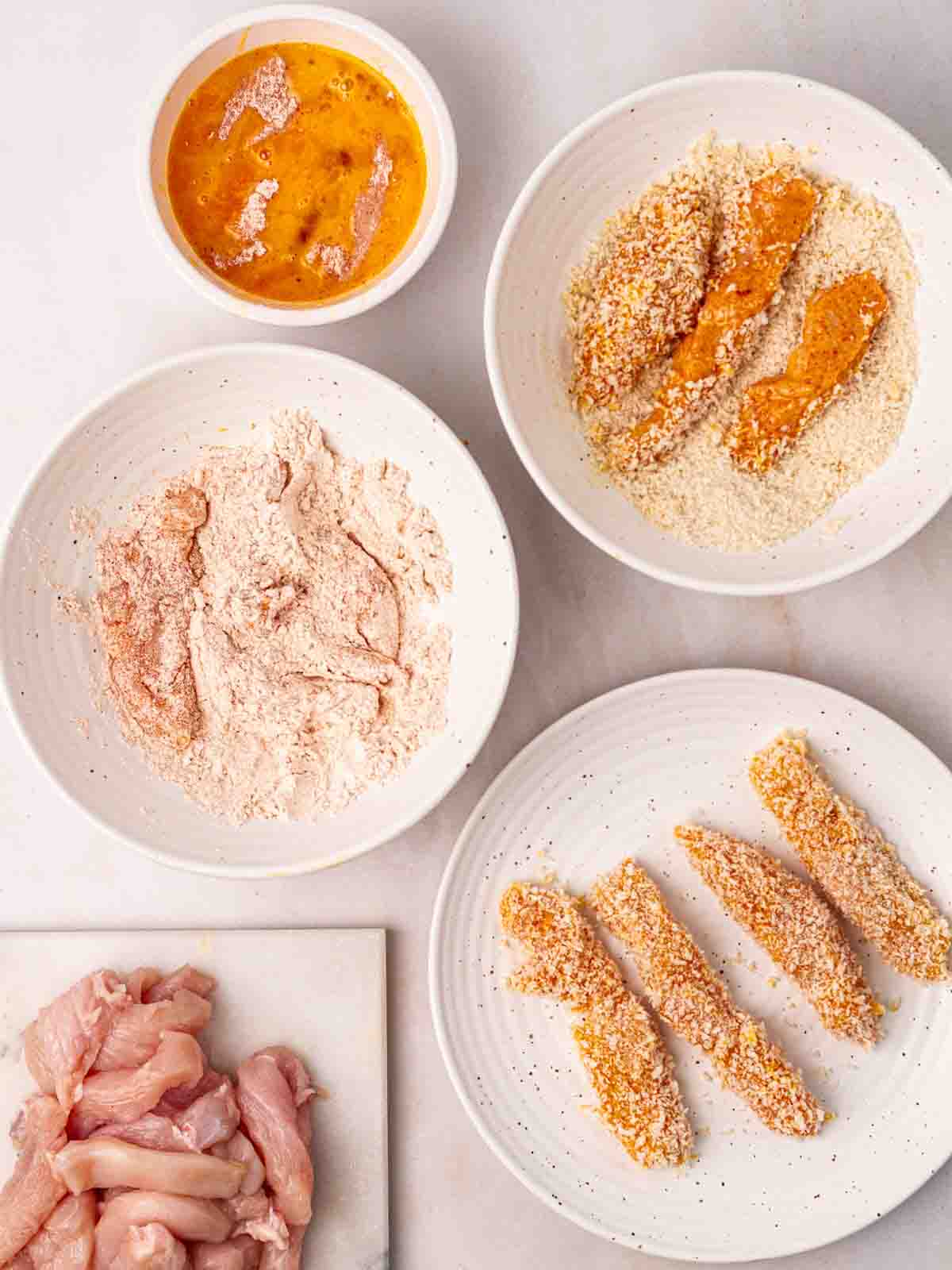 Several bowls of ingredients to coat pieces of raw chicken in breadcrumbs to make homemade goujons.