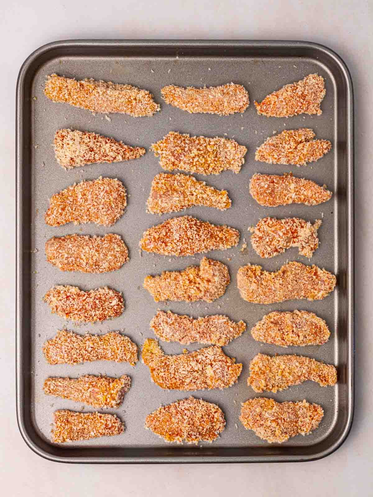 Raw chicken goujons on a baking tray coated in breadcrumbs, ready for oven baking.
