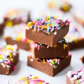 Three squares of chocolate fudge with sprinkles stacked up on a table, with other pieces around the sides.