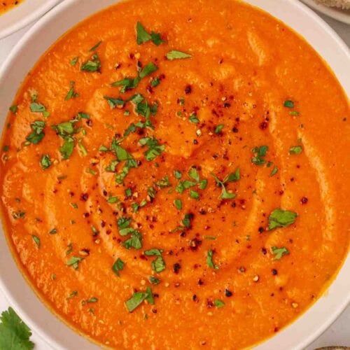 A bowl of orange soup made with carrots and lentils and pepper on top, with crusty bread to the side.