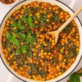 A large white pan filled with Chickpea Curry with spinach and mint garnish, and a wooden spoon stirring.