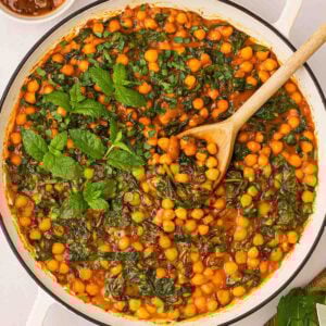 A large white pan filled with Chickpea Curry with spinach and mint garnish, and a wooden spoon stirring.