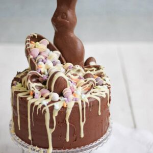A very chocolatey Easter chocolate cake with lots of chocolate egg and bunny toppings and melted white chocolate, on a cake stand.