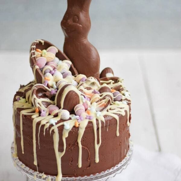 A very chocolatey Easter chocolate cake with lots of chocolate egg and bunny toppings and melted white chocolate, on a cake stand.