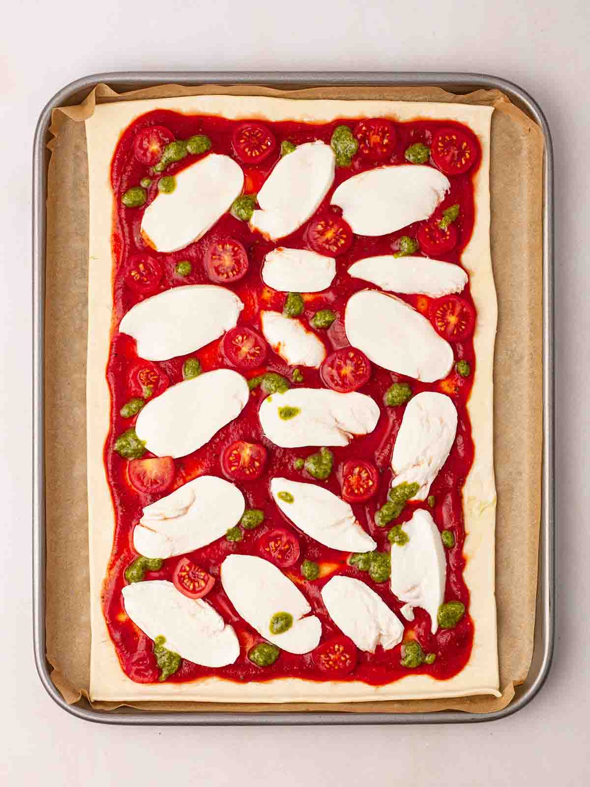 An uncooked Puff Pastry Pizza on a baking tray with baking paper, topped with tomato sauce, mozzarella, tomatoes and pesto.