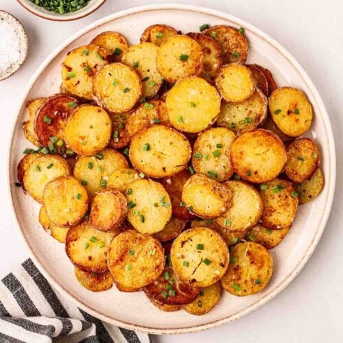 A plate of potatoes that have been sautéed on a white table with chives in a bowl to the side and a stripy tea towel nearby.