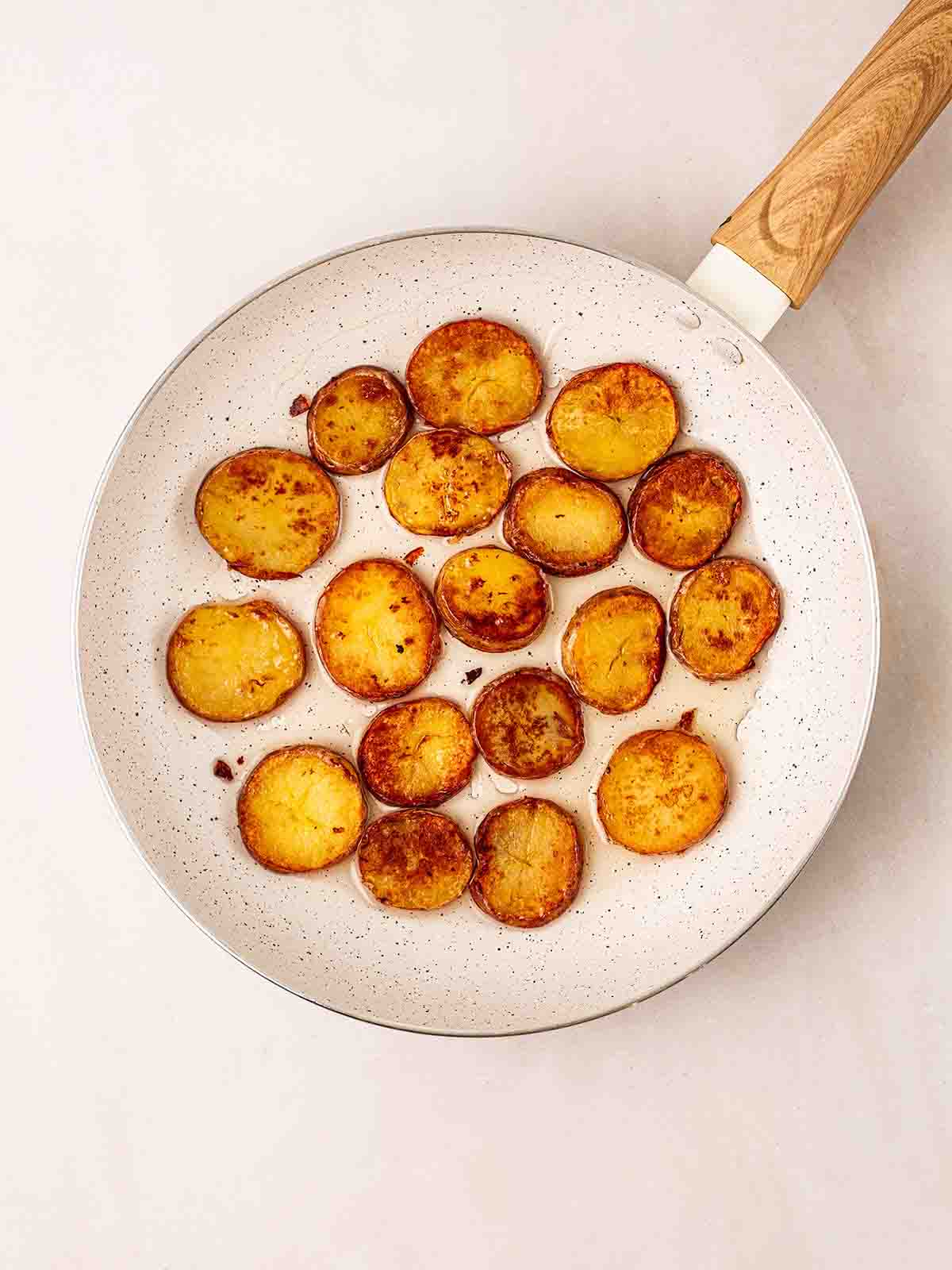 A white pan filled with a single layer of sliced potatoes frying in oil, going golden. For step 2 in the recipe for Sautéed Potatoes.