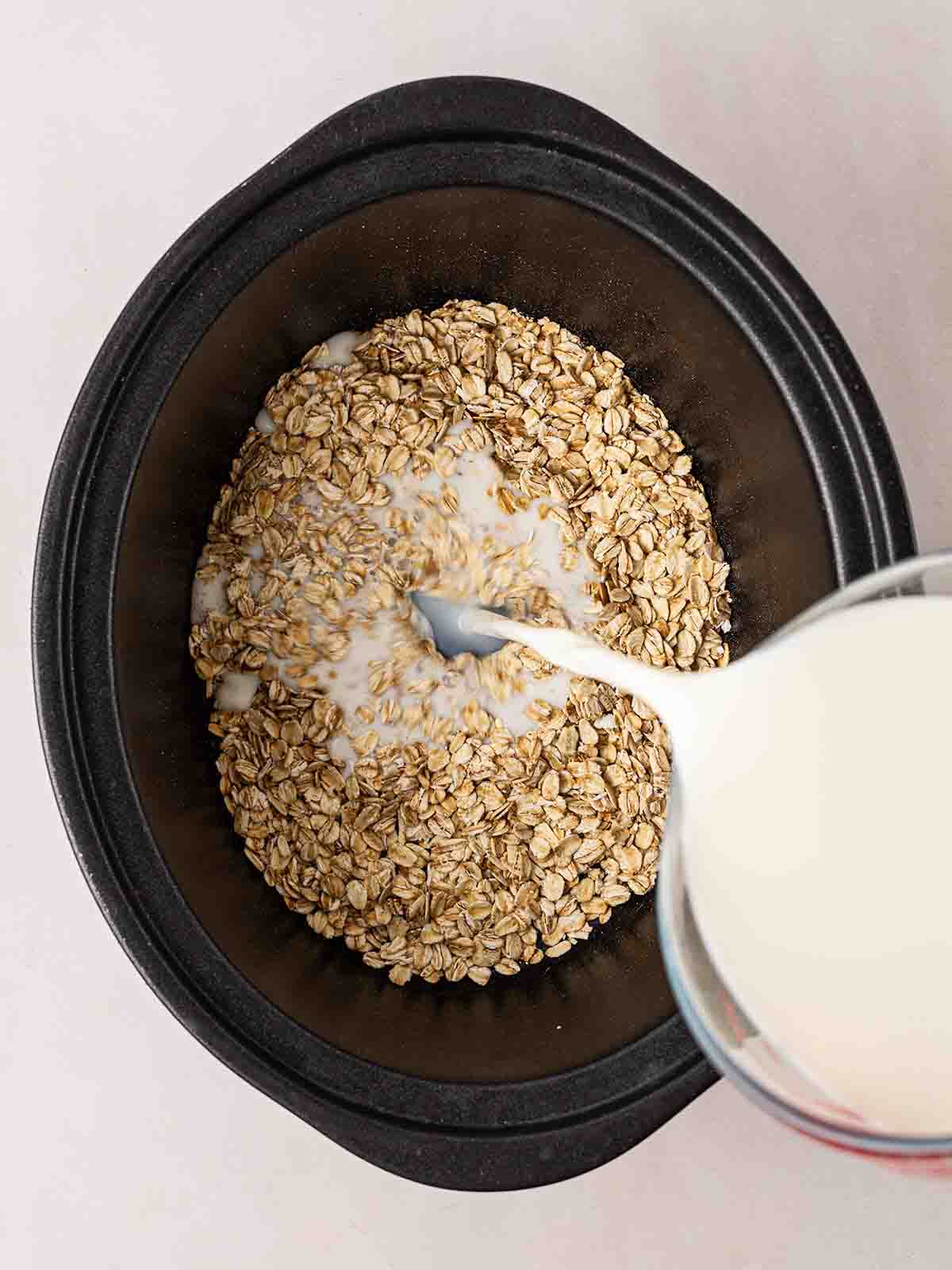 A slow cooker pan filled with oats, with milk being poured in, ready to make Slow Cooker Porridge.